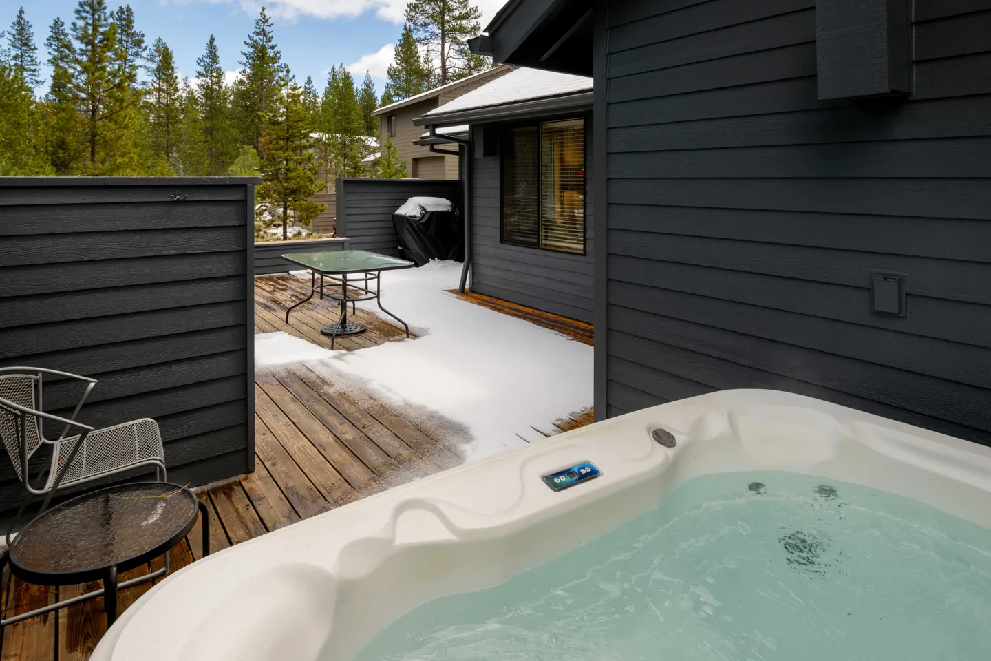 Outdoor hot tub filled with clear water on a wooden deck with snow patches, dark siding, and patio furniture.