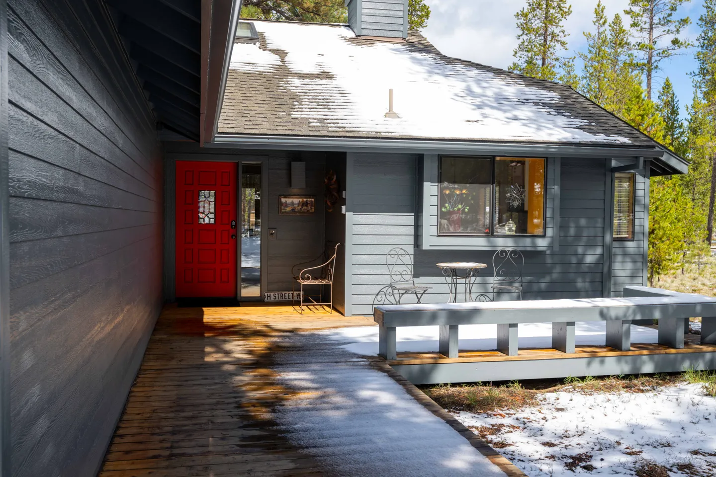Front porch and entryway of a gray house with a bright red front door, snow on the ground and roof, and a wooden deck with seating.