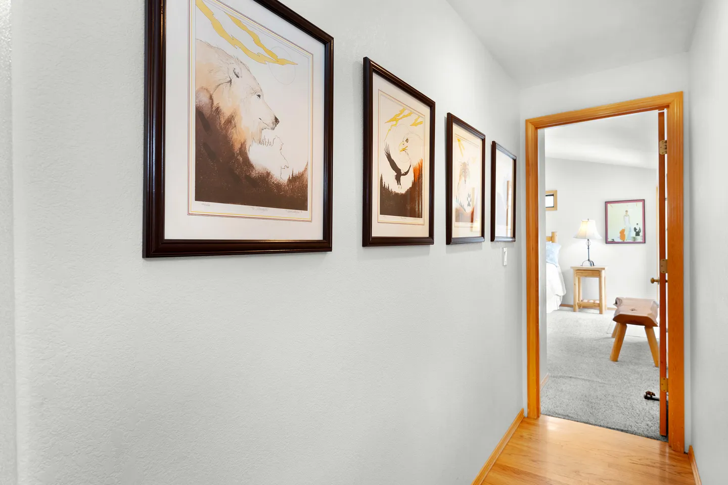 Hallway lined with framed art on a white wall, looking into a carpeted bedroom through a wood-trimmed doorway.