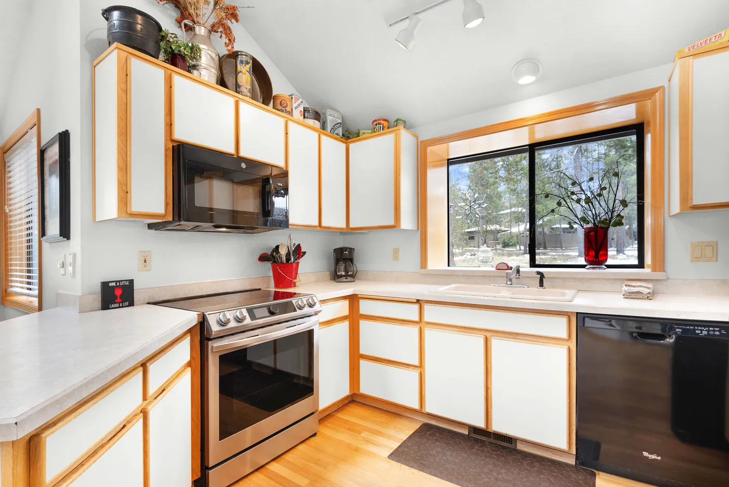 Kitchen interior featuring white cabinets with light wood trim, stainless steel oven, black microwave, and a window above the sink.