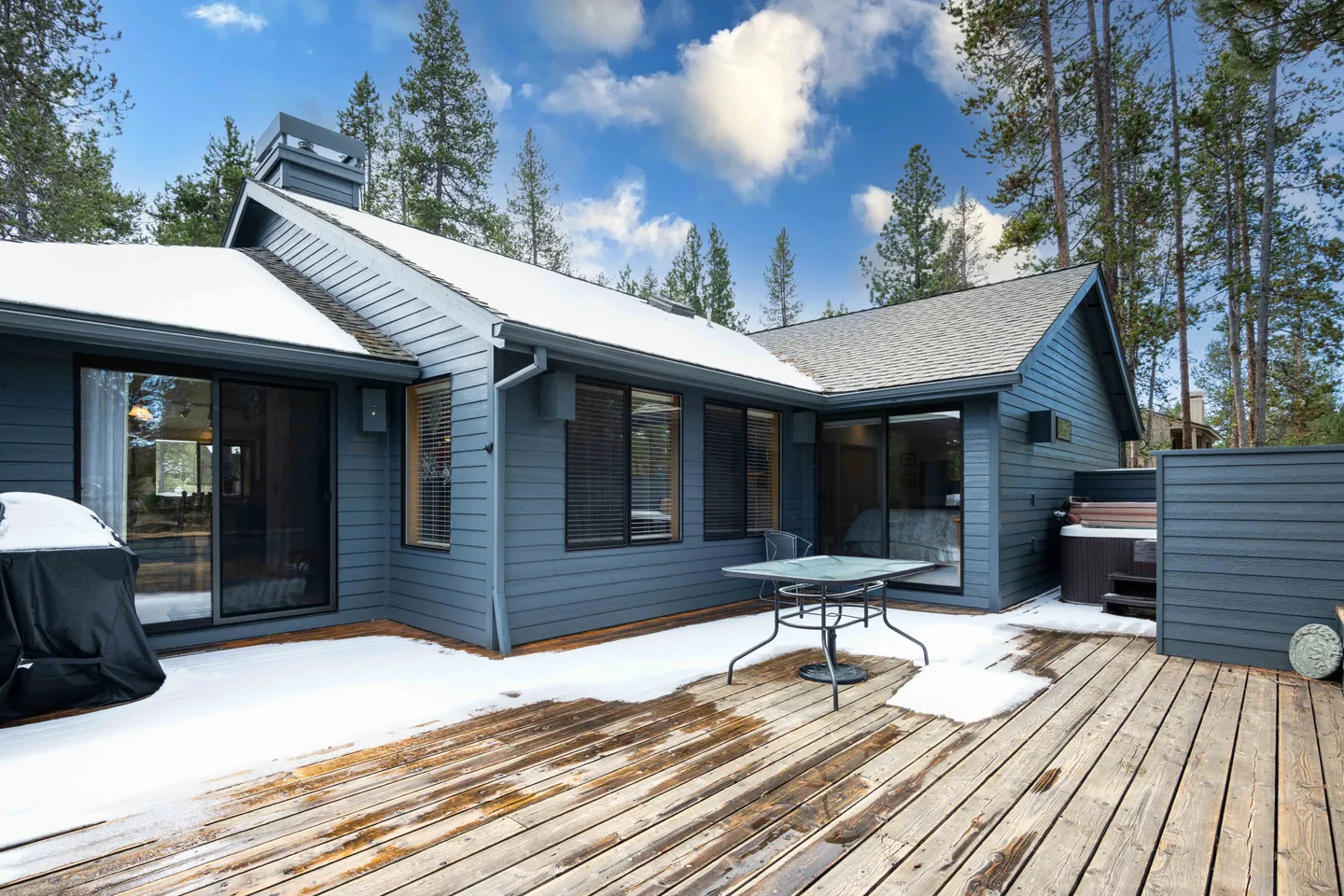 Rear wooden deck of a blue-gray home with patches of snow, a small outdoor table, a covered grill, and a hot tub.