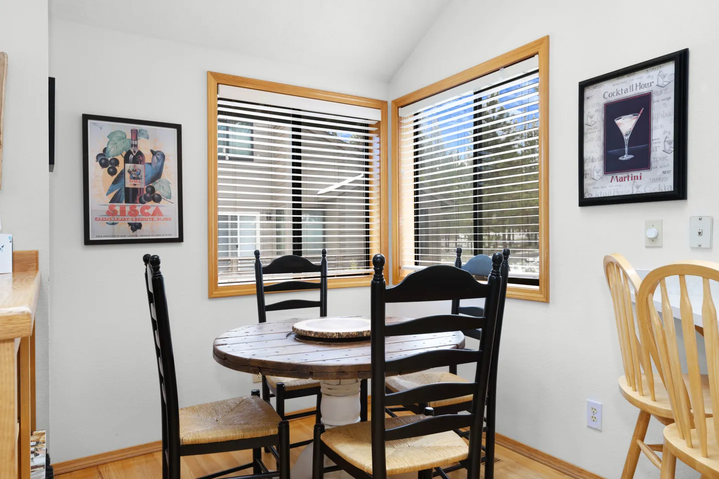 Dining nook with a round wooden table, black ladder-back chairs, and two corner windows with blinds.