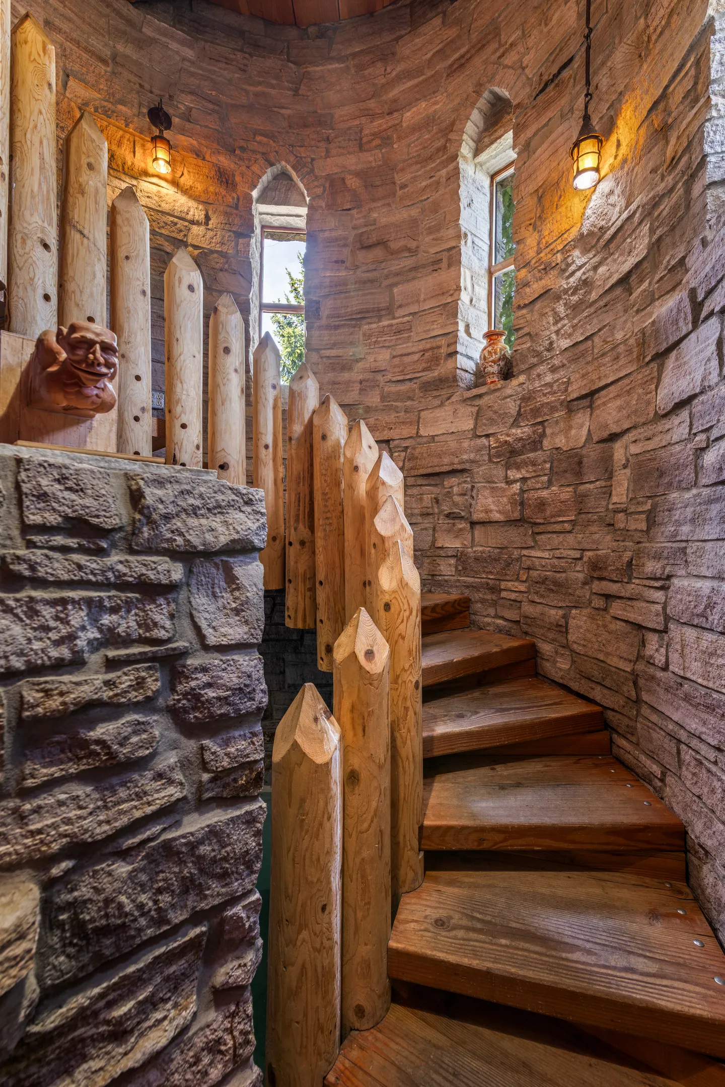 A rustic stone turret featuring a spiral staircase with wood steps, pointed log posts, arched windows, and lantern light fixtures.