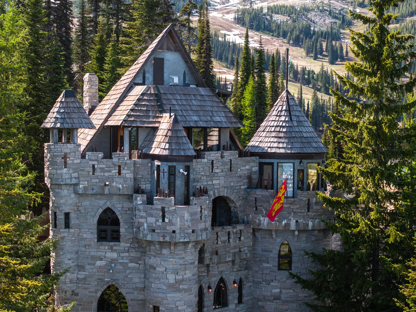 Exterior of a castle-style home with stone walls, multiple turrets, and shingle roofs set among pine trees with mountains in the background.