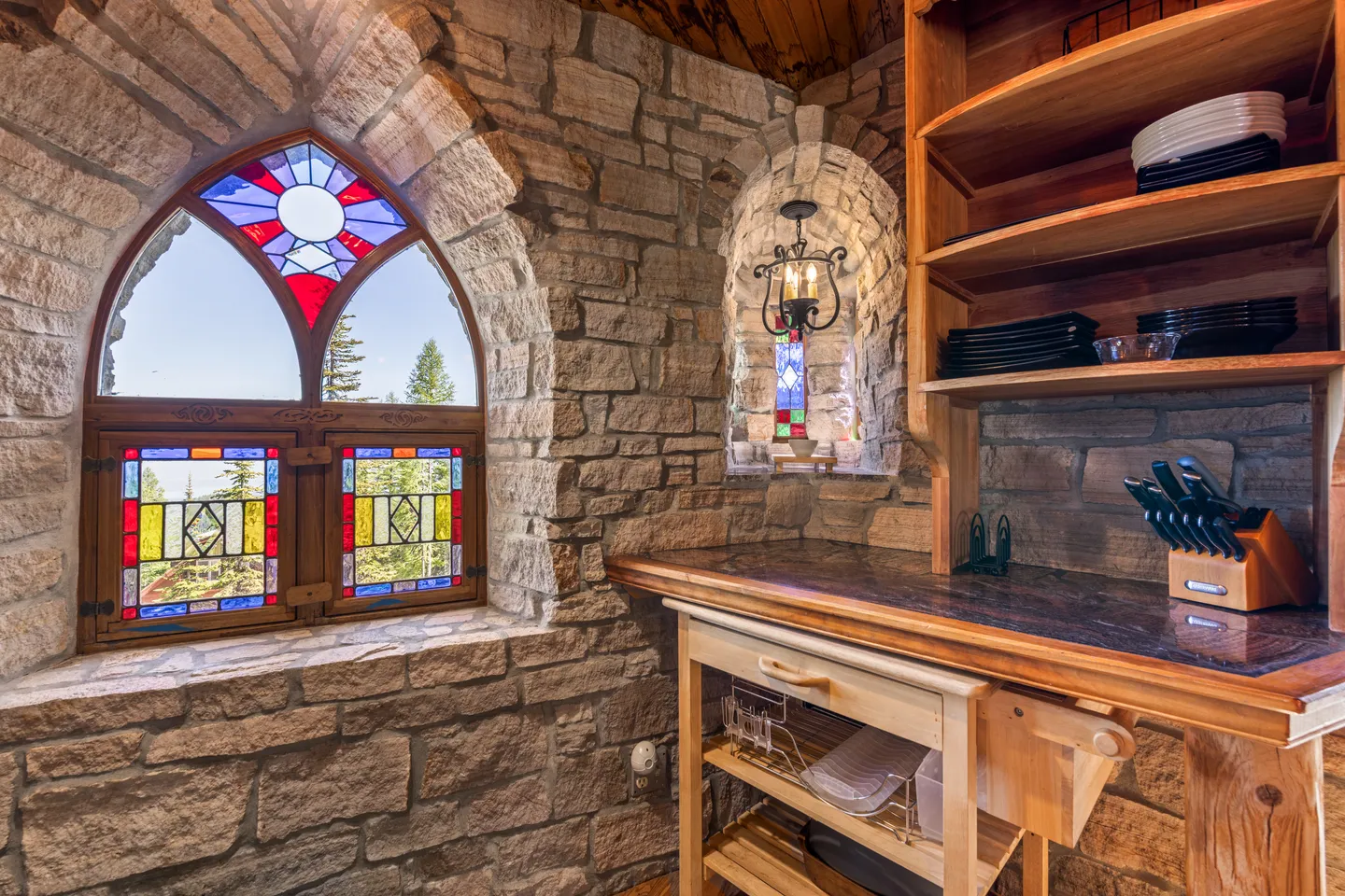 Stone alcove interior featuring an arched window with stained glass panes, a smaller lit niche, and a wooden prep cart beneath shelving.