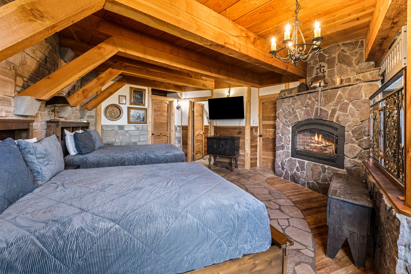 Rustic bedroom with exposed wood ceiling beams, two beds with blue comforters, and a lit stone fireplace.