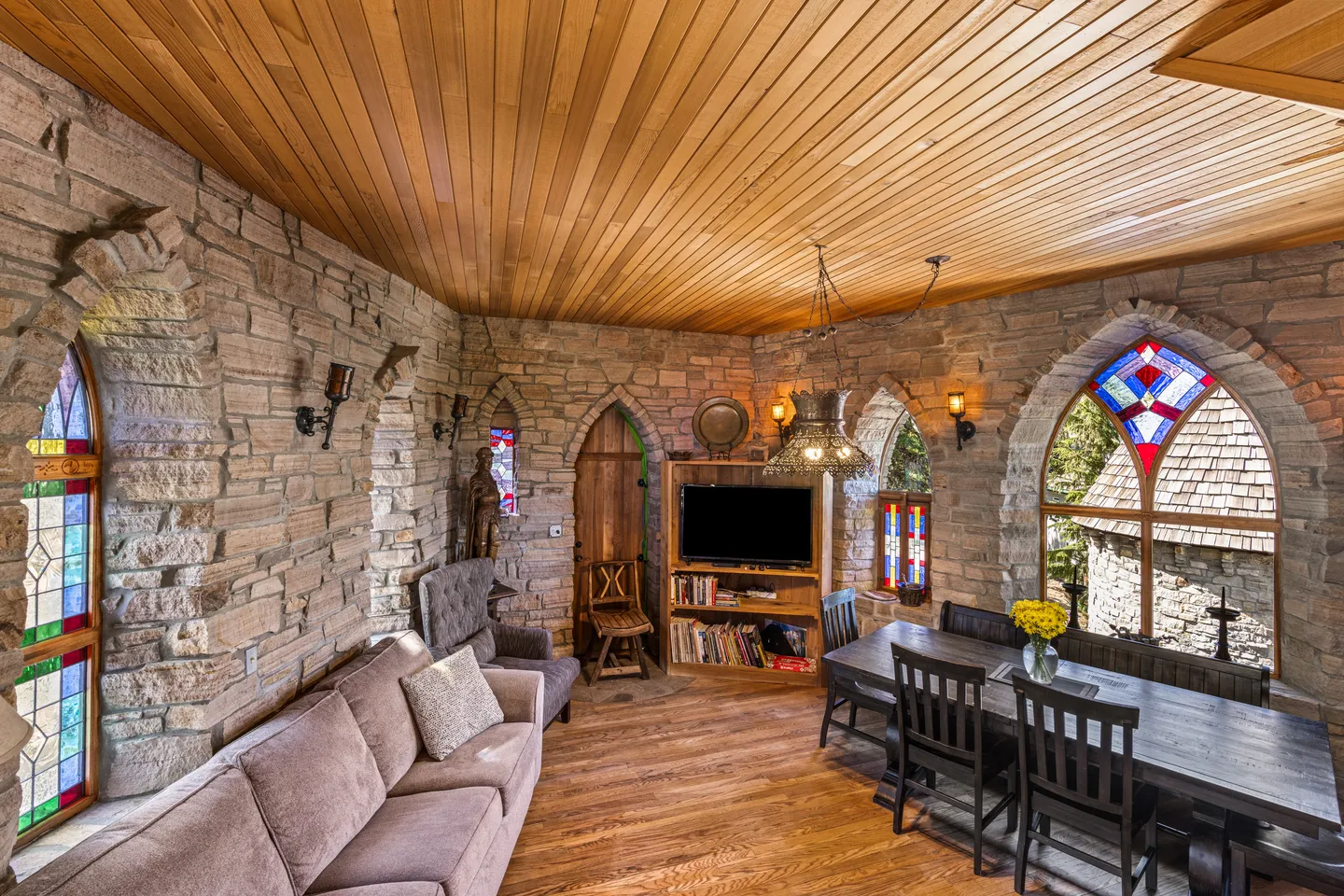 Living and dining space with stone walls, arched stained glass windows, wood ceiling, sofa, TV unit, and dining table.