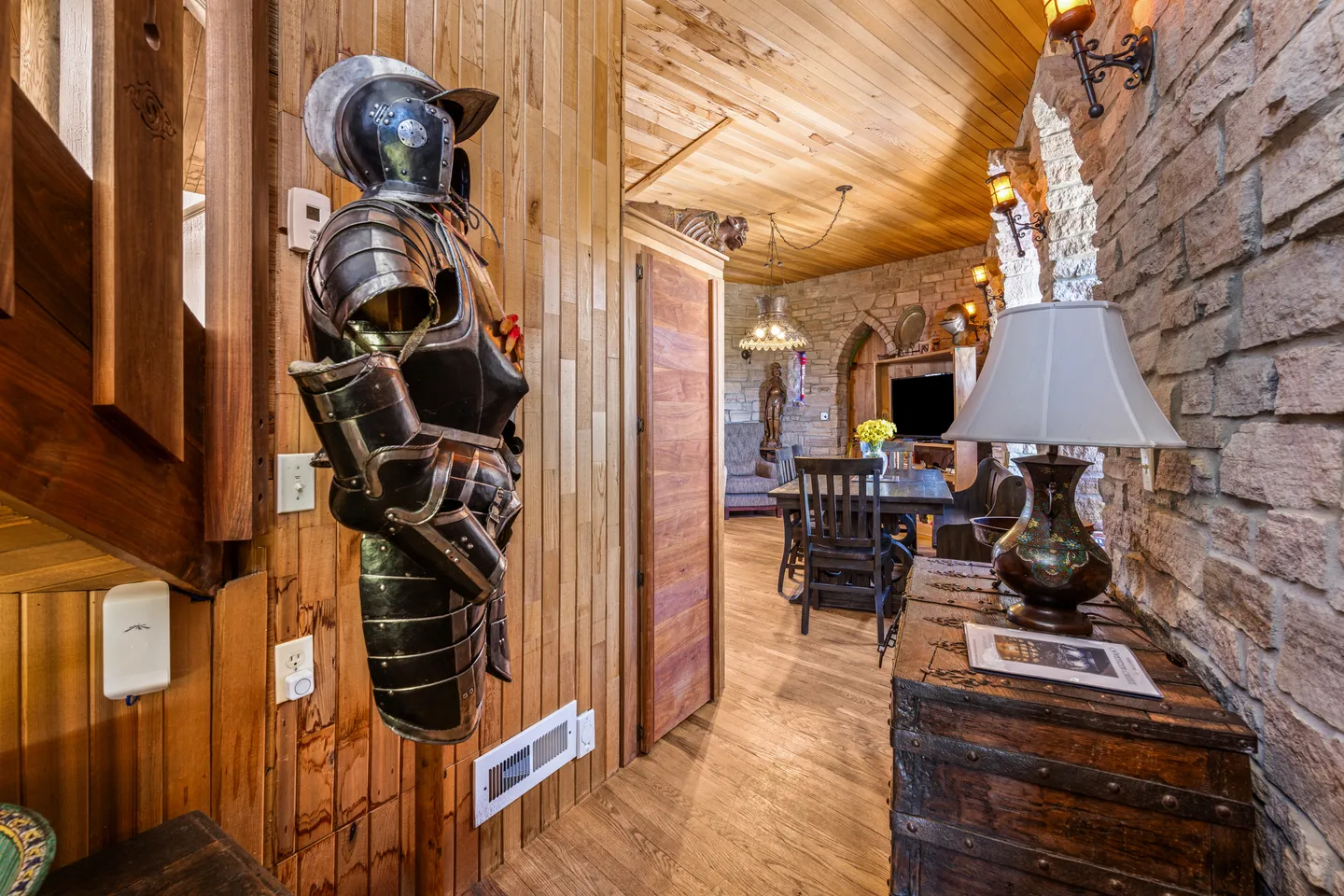 Hallway featuring wood paneling, a suit of armor display, stone walls, and a view into a dining area.