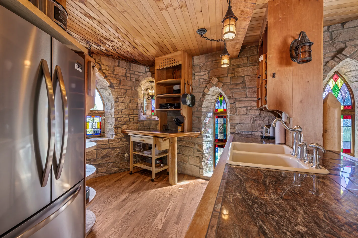 A kitchen with stone walls, wood plank ceiling, stainless steel refrigerator, and arched windows with stained glass.