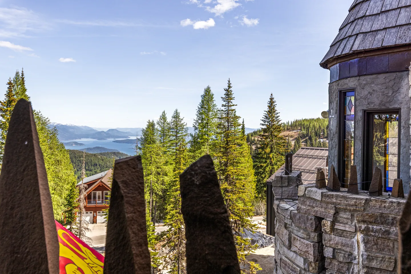 Elevated view through stone castle-style battlements overlooking evergreen trees, surrounding homes, and a distant lake under a blue sky.