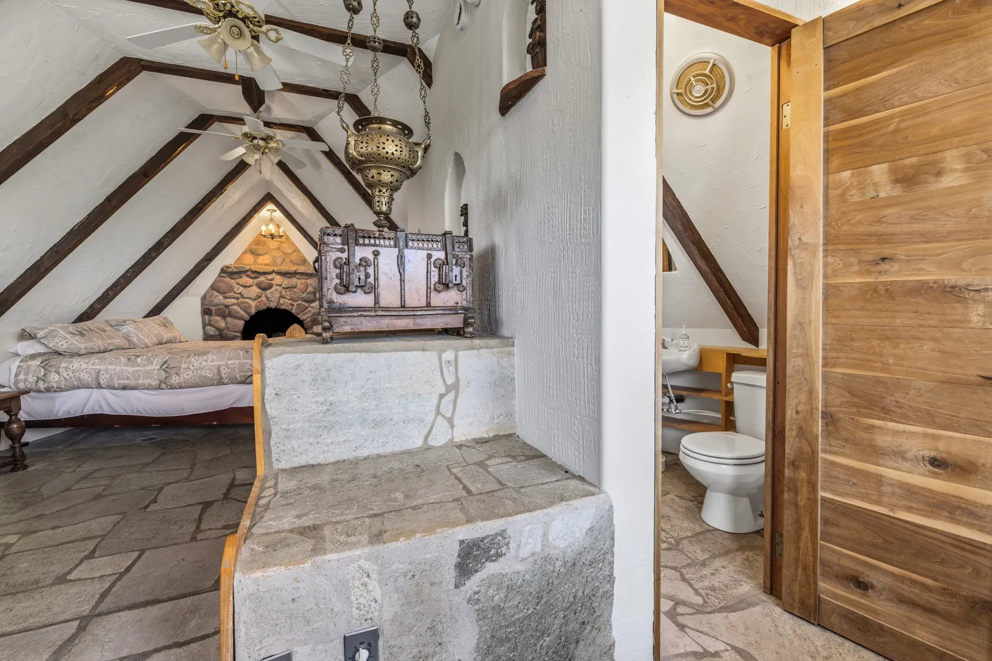 A rustic bedroom with stone floors, pitched ceiling with exposed wood beams, and a stone fireplace, visible past an open bathroom doorway.