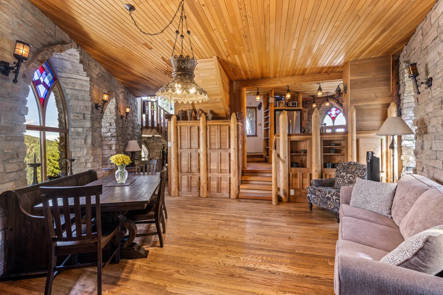 Open-concept interior featuring stone walls with arched stained glass windows, wood-paneled ceilings, a dining table, and living room seating.