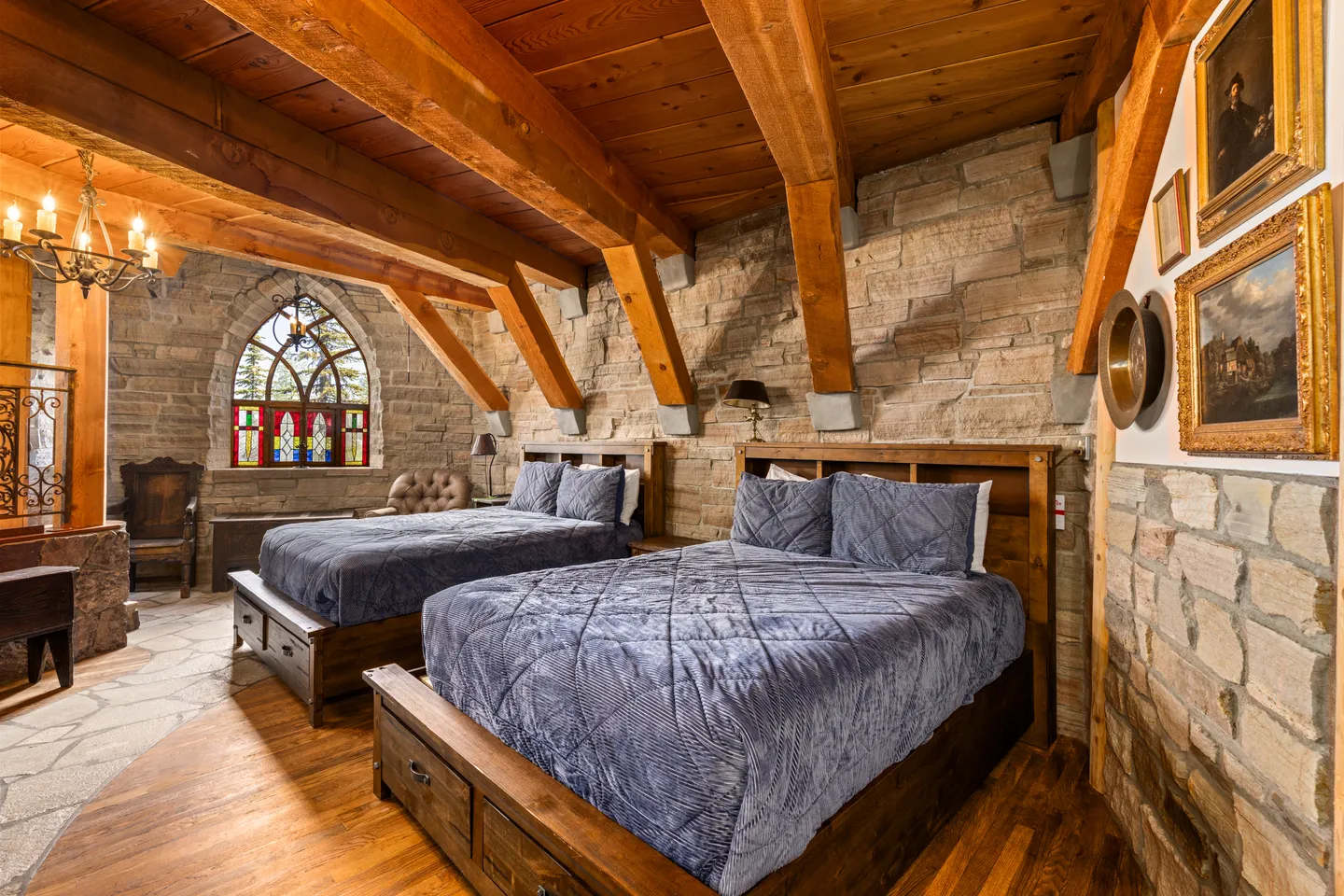 Bedroom featuring stone walls, exposed timber ceiling beams, two wood-framed beds, and a stained glass gothic window.