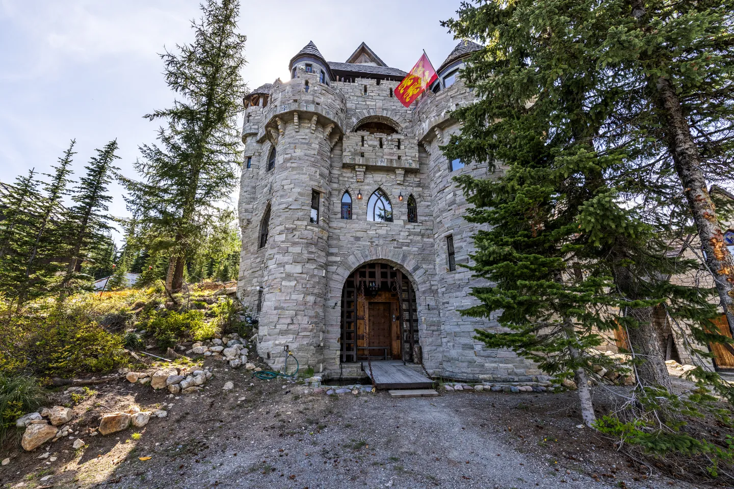 Exterior low-angle view of a multi-story stone castle-style building with turrets, arched entrance, and surrounding pine trees.