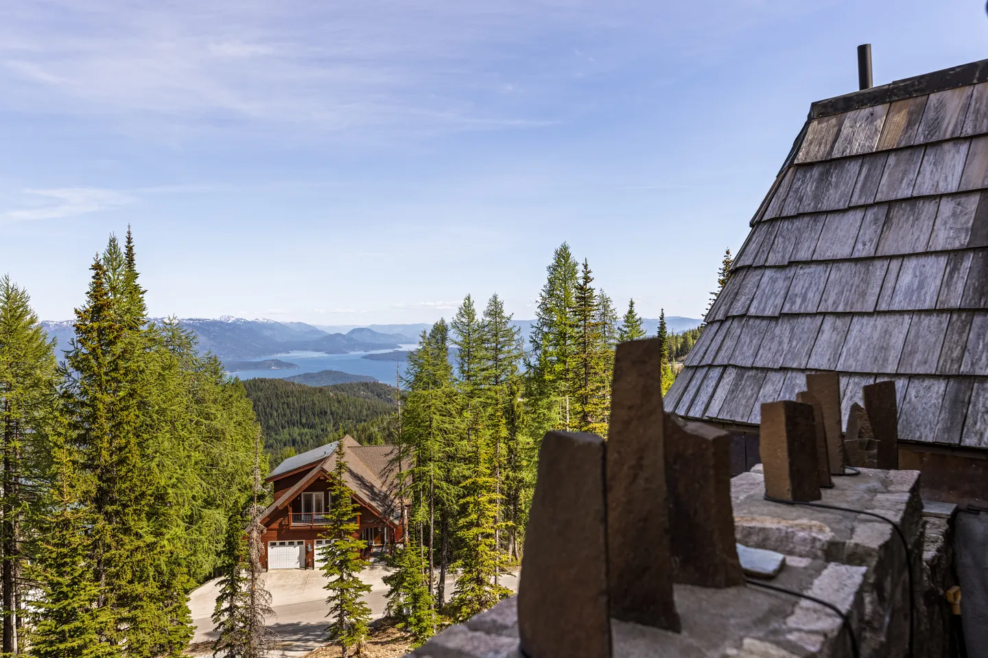 Elevated outdoor view from a stone balcony overlooking tall evergreen trees, a neighboring house, and a distant lake and mountains under a blue sky.