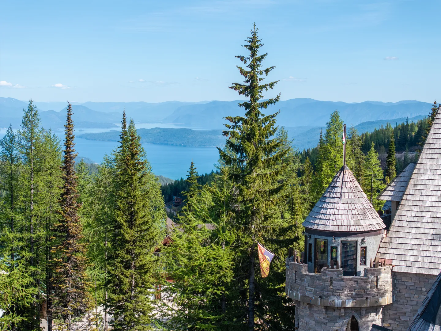 Elevated view of a stone castle tower overlooking evergreen trees and a large blue lake surrounded by mountains.
