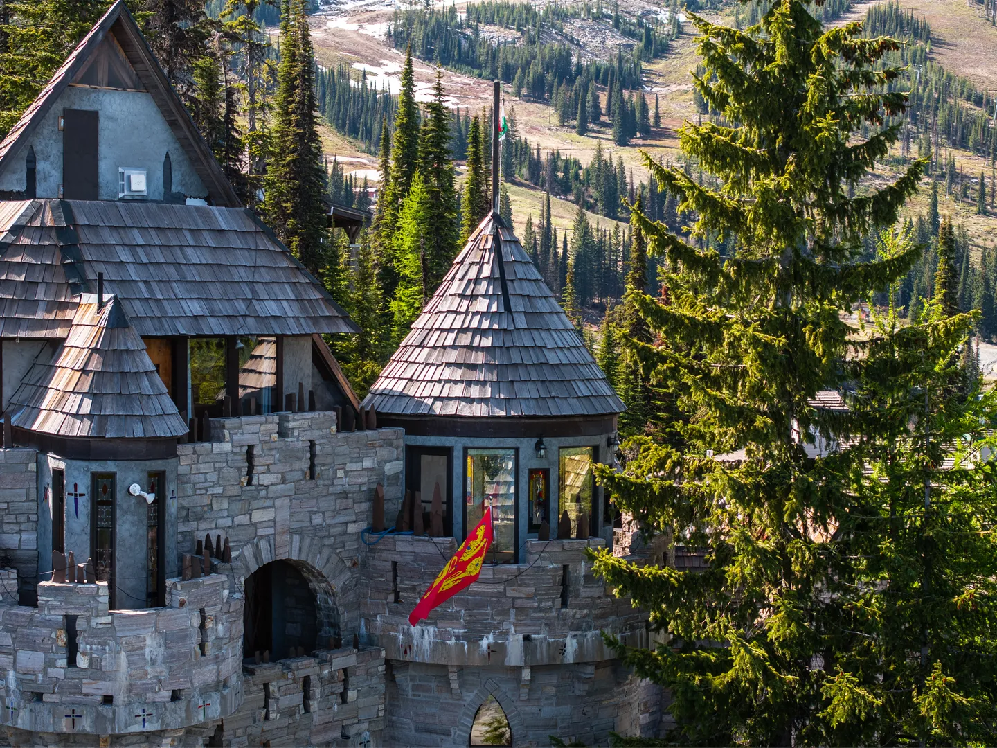 Exterior of a castle-style home with stone walls, shingled conical turret, and battlements set against a pine-covered hillside.
