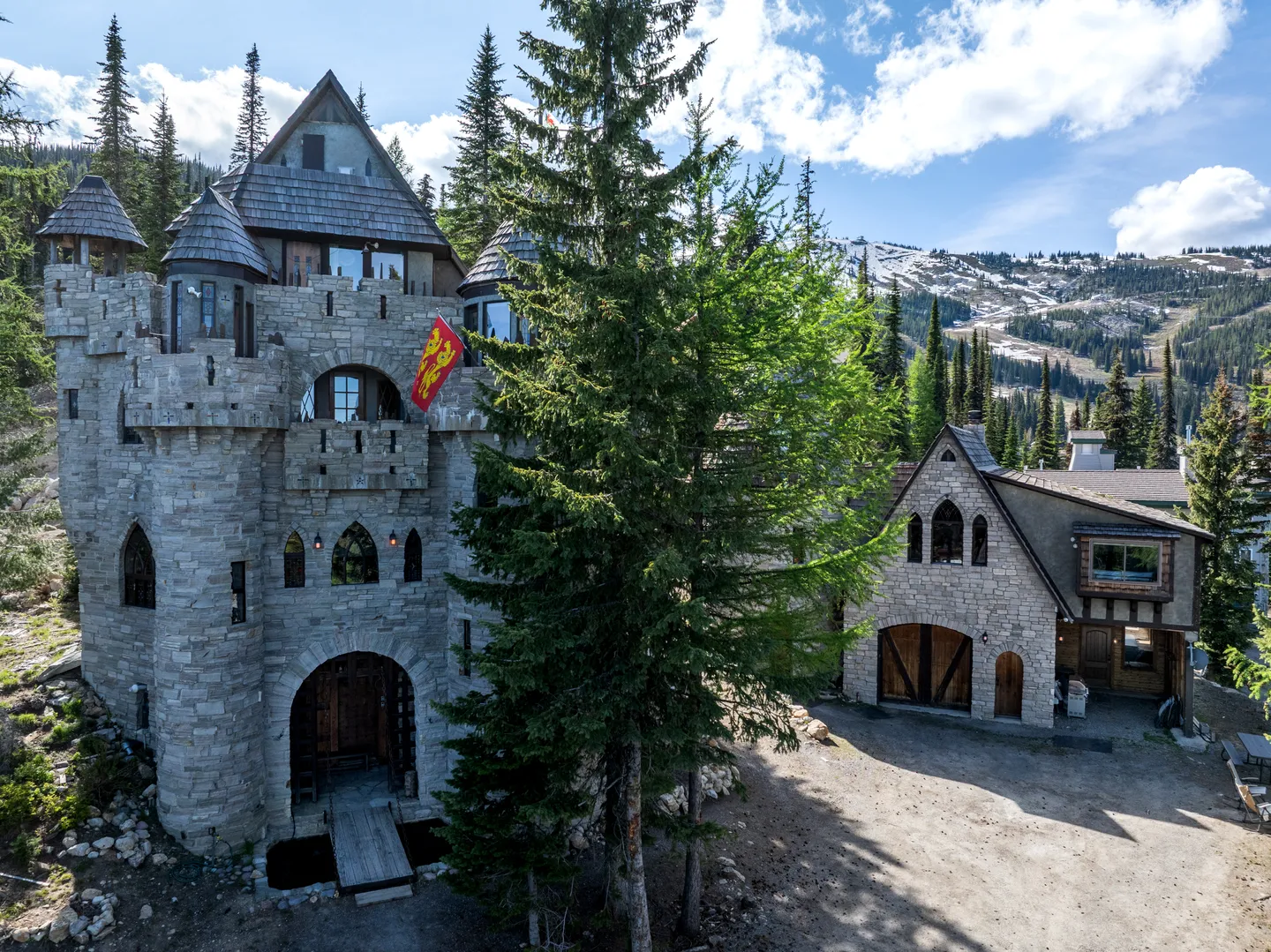 Exterior of a multi-story stone castle-style house with turrets, an arched entryway, and surrounding pine trees against mountain slopes.
