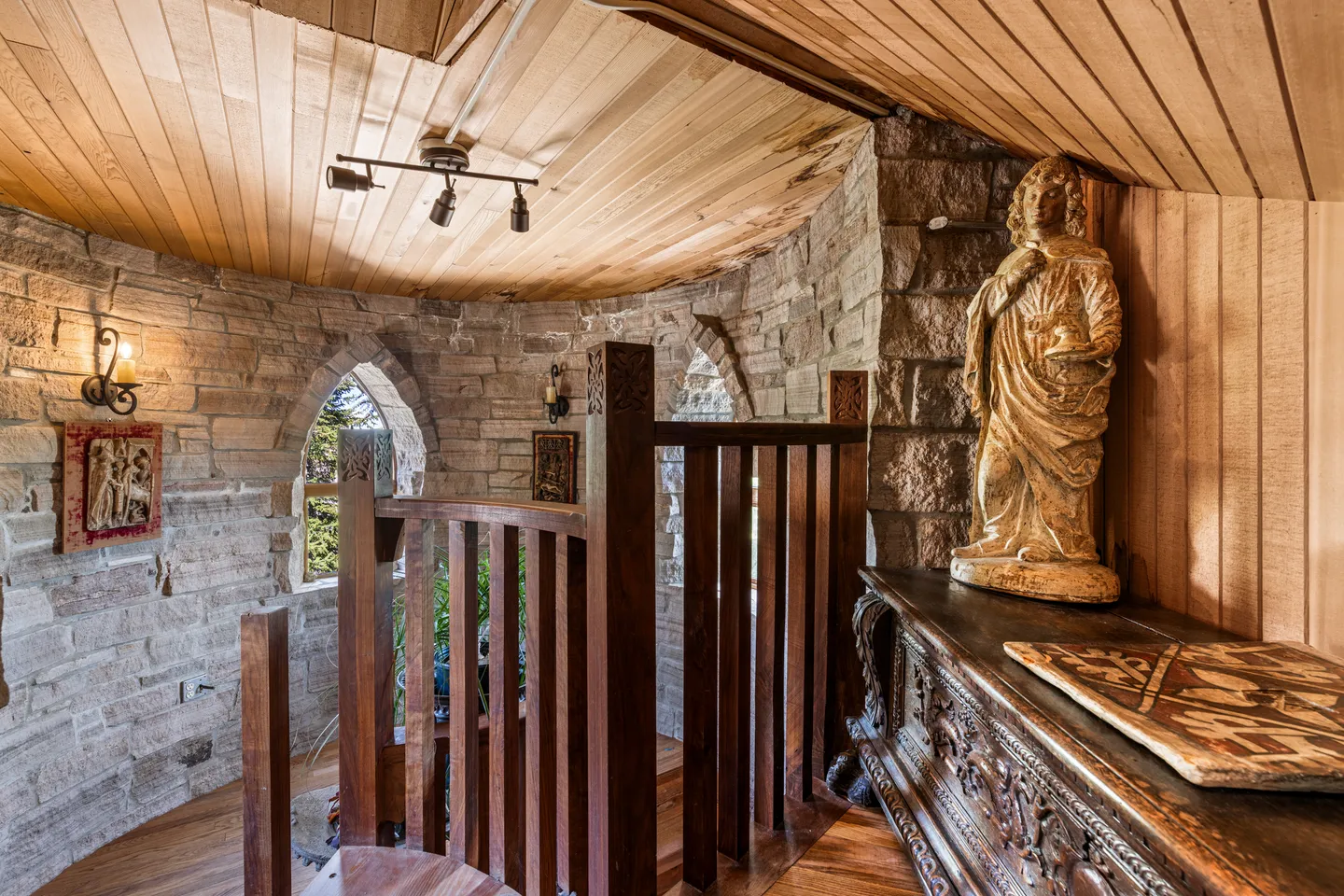 Interior view of an upper landing with stone walls, arched windows, wood-plank ceiling, and a wooden staircase railing.