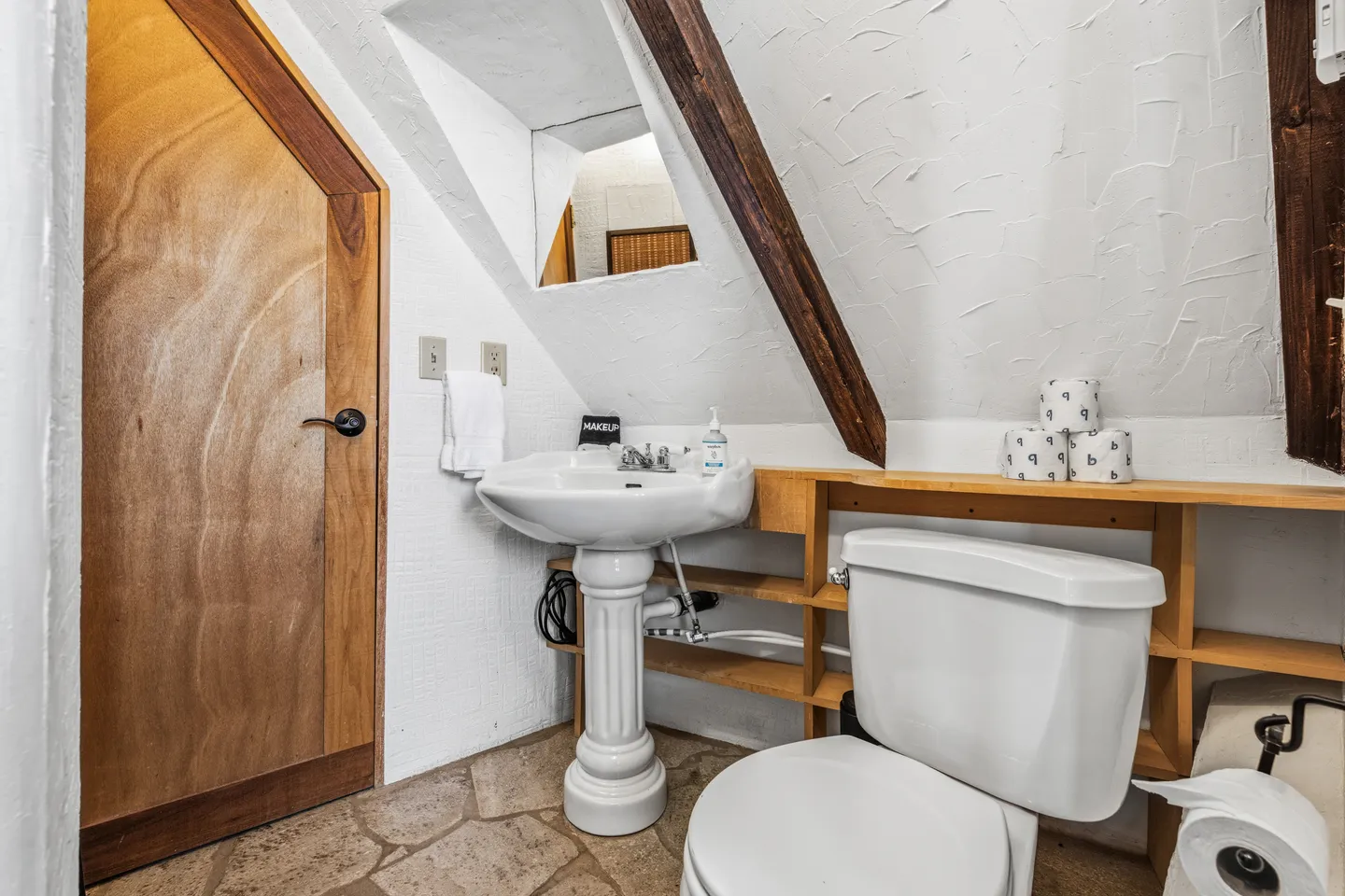 Bathroom with sloped ceiling, exposed wood beam, pedestal sink, toilet, and uniquely shaped wooden door.