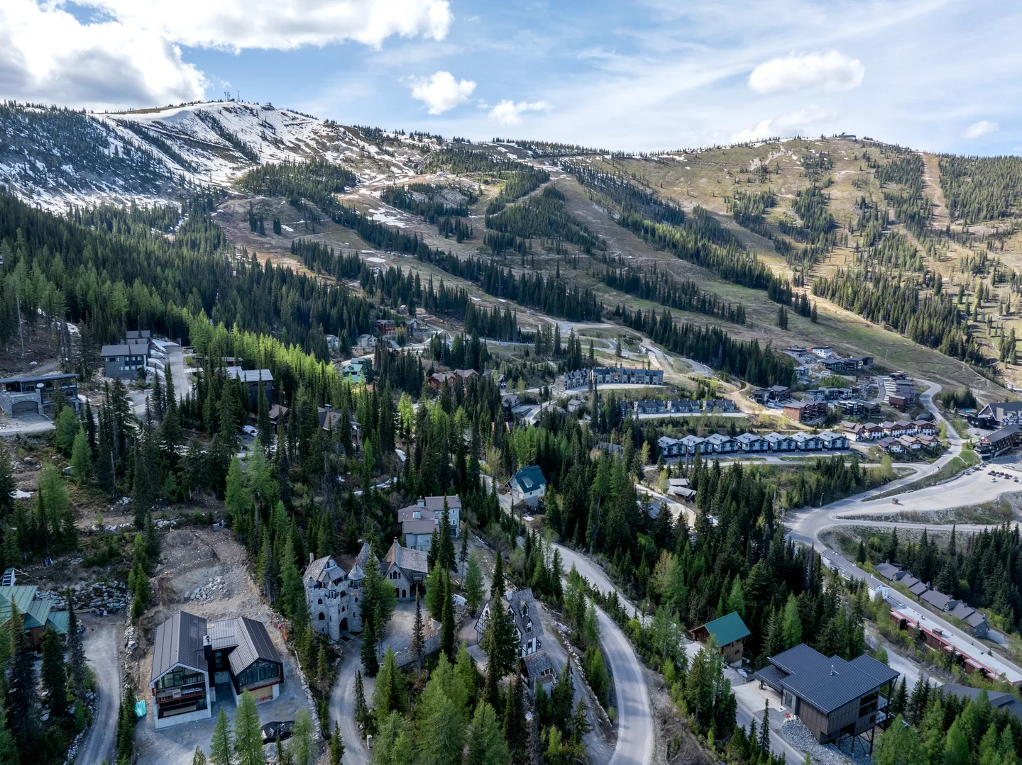 Aerial view of a mountain resort community nestled in evergreen trees with ski slopes and snow-capped peaks in the background.