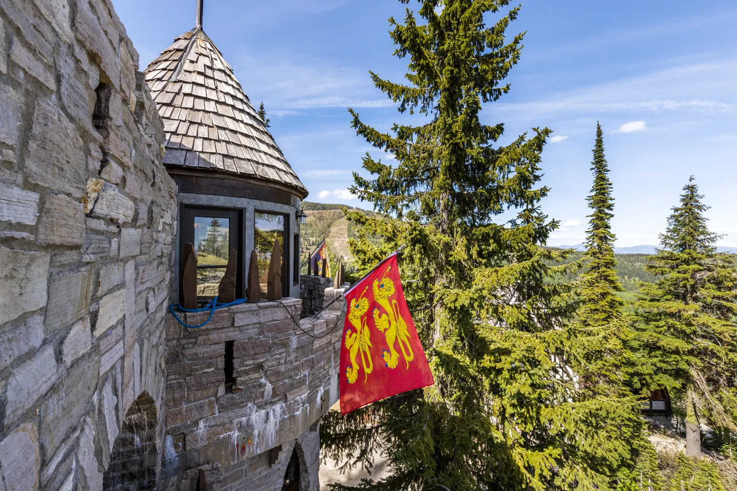 Exterior stone turret and balcony of a castle-style building featuring a red flag with yellow heraldic lions, surrounded by tall pine trees.