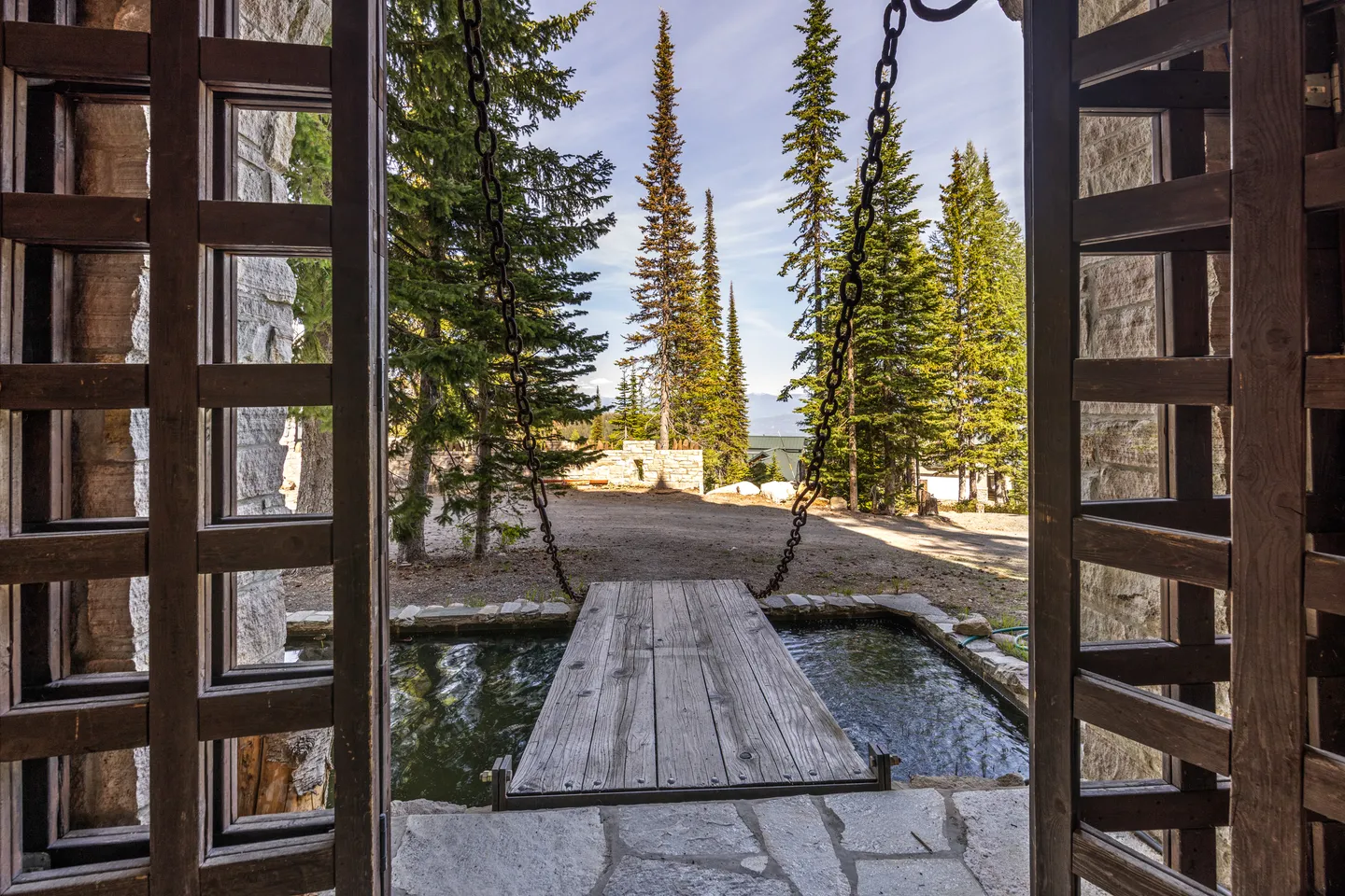 A view looking out an open wooden entryway across a lowered wooden drawbridge spanning a small moat toward pine trees.