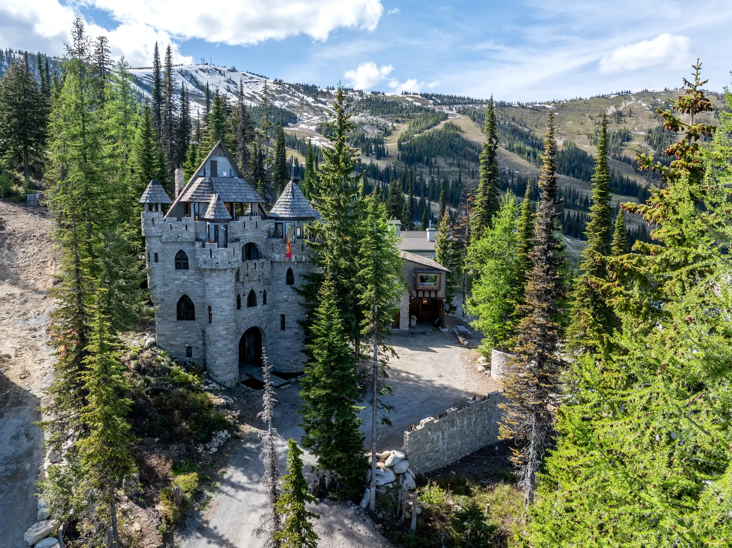 Aerial view of a stone castle-style house surrounded by tall pine trees with ski slopes and mountains in the background.