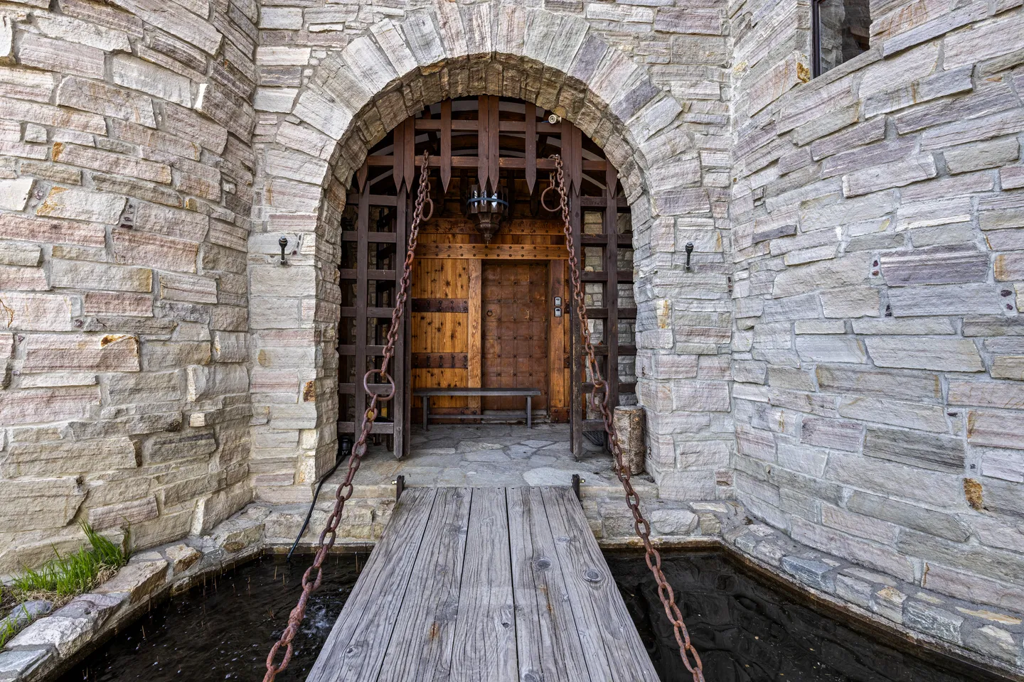A stone castle-style entrance featuring a wooden bridge flanked by chains over a moat, leading to an arched doorway with a wooden portcullis.