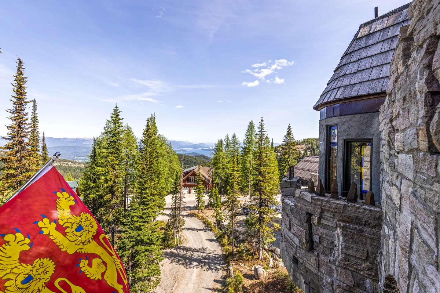 Elevated view from a stone castle-style tower overlooking a gravel driveway, evergreen trees, and distant mountains under a blue sky.