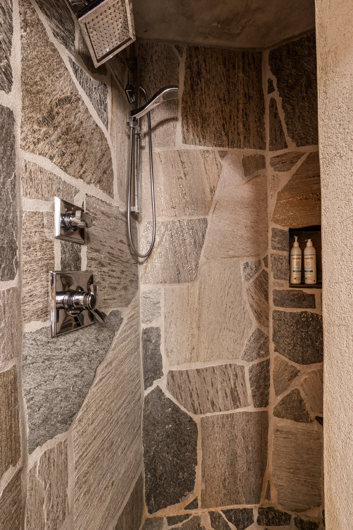 A walk-in shower with natural stone flagstone walls, chrome fixtures, a rain showerhead, and a built-in niche with toiletries.