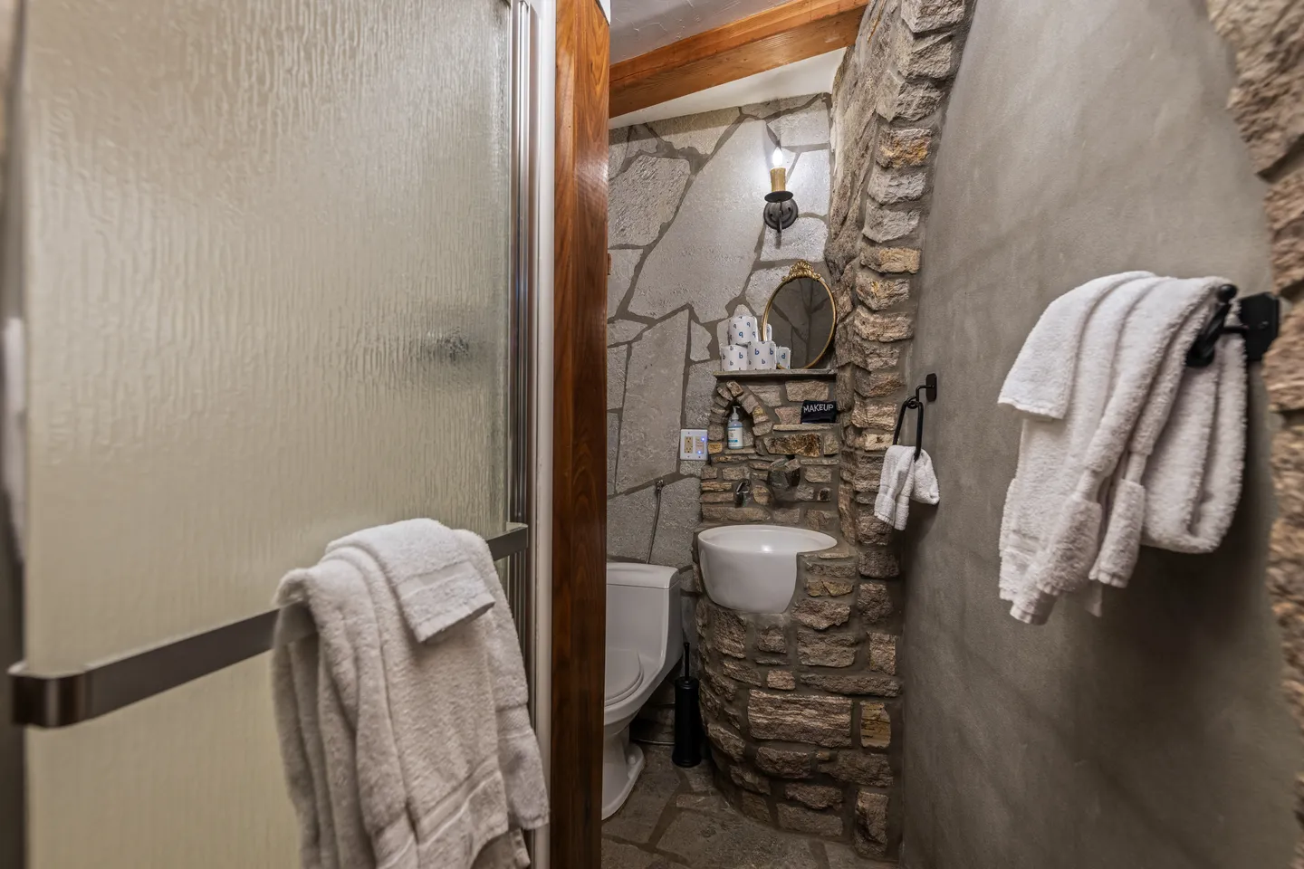 Bathroom featuring stone masonry walls, a small corner sink, a toilet, and textured glass shower doors with towels.