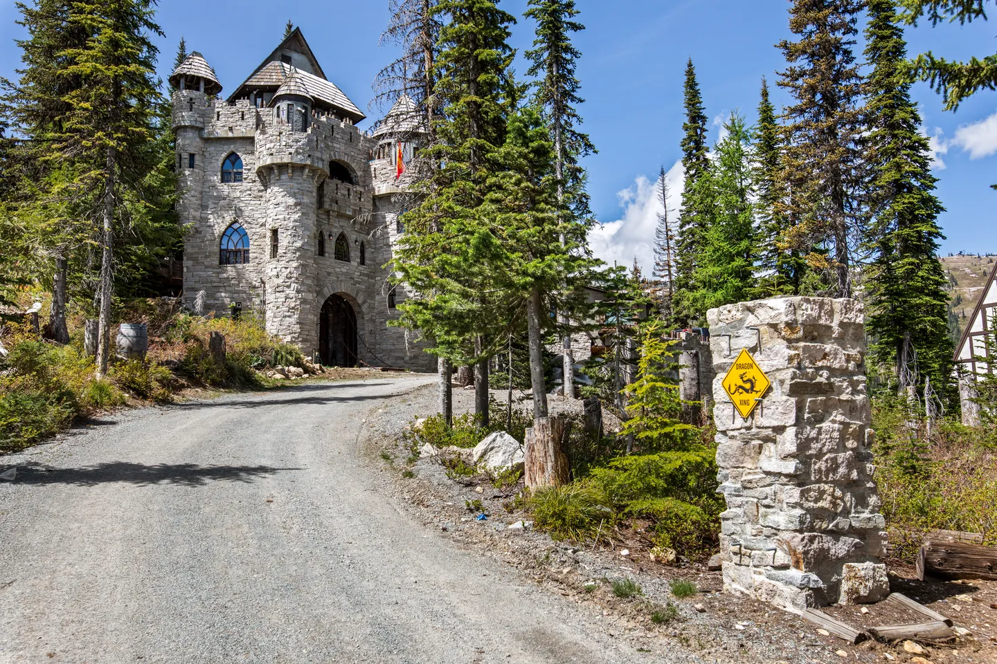 A gravel driveway leads to a stone castle-style structure surrounded by evergreen trees under a bright blue sky.