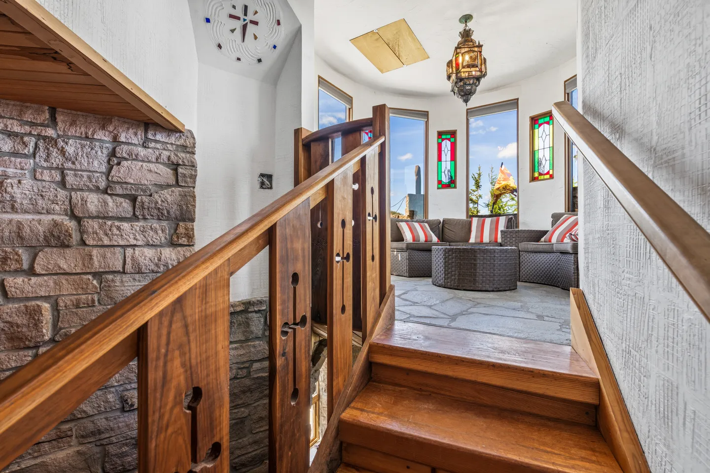 A wooden staircase leads up to a curved sunroom with stone flooring, woven wicker seating, and stained-glass accent windows.