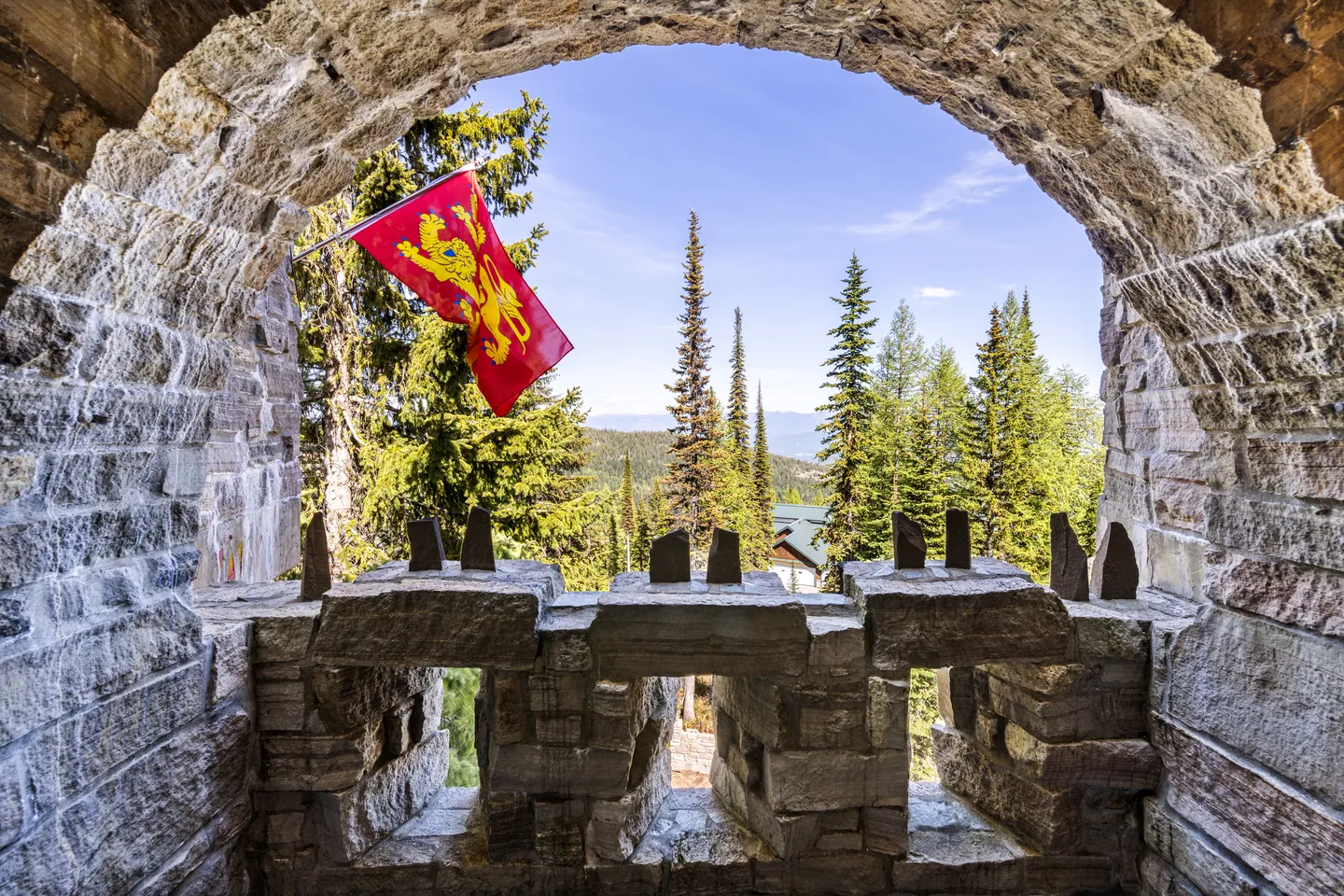 View of evergreen trees and mountains framed through an arched stone turret opening with a red lion flag.