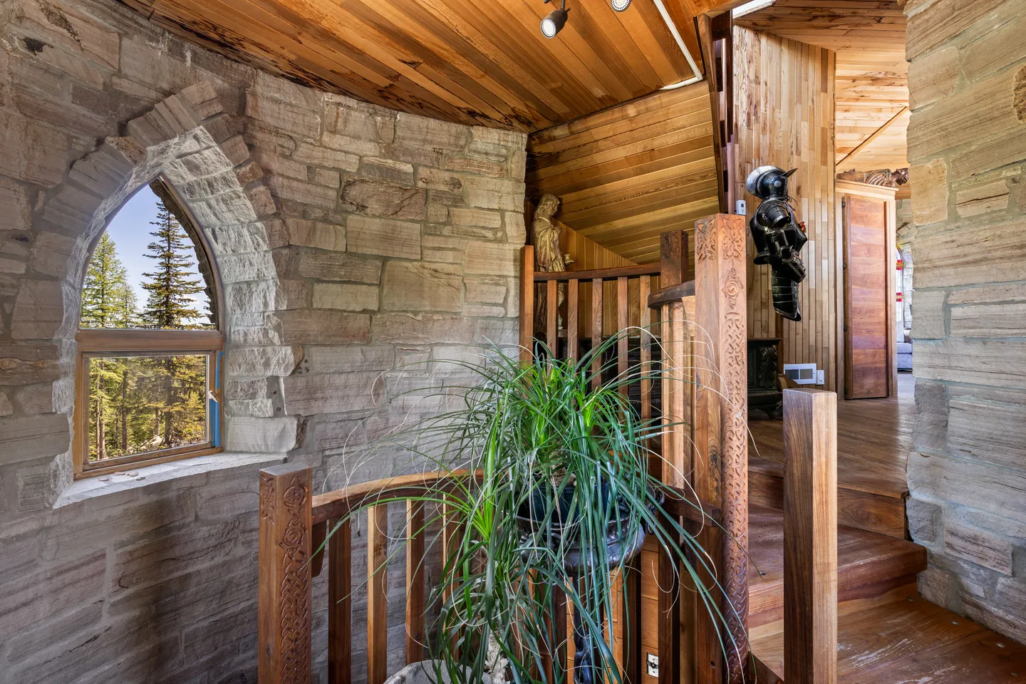 A rustic indoor staircase landing featuring natural stone walls, wood plank ceilings, and an arched window looking out at pine trees.
