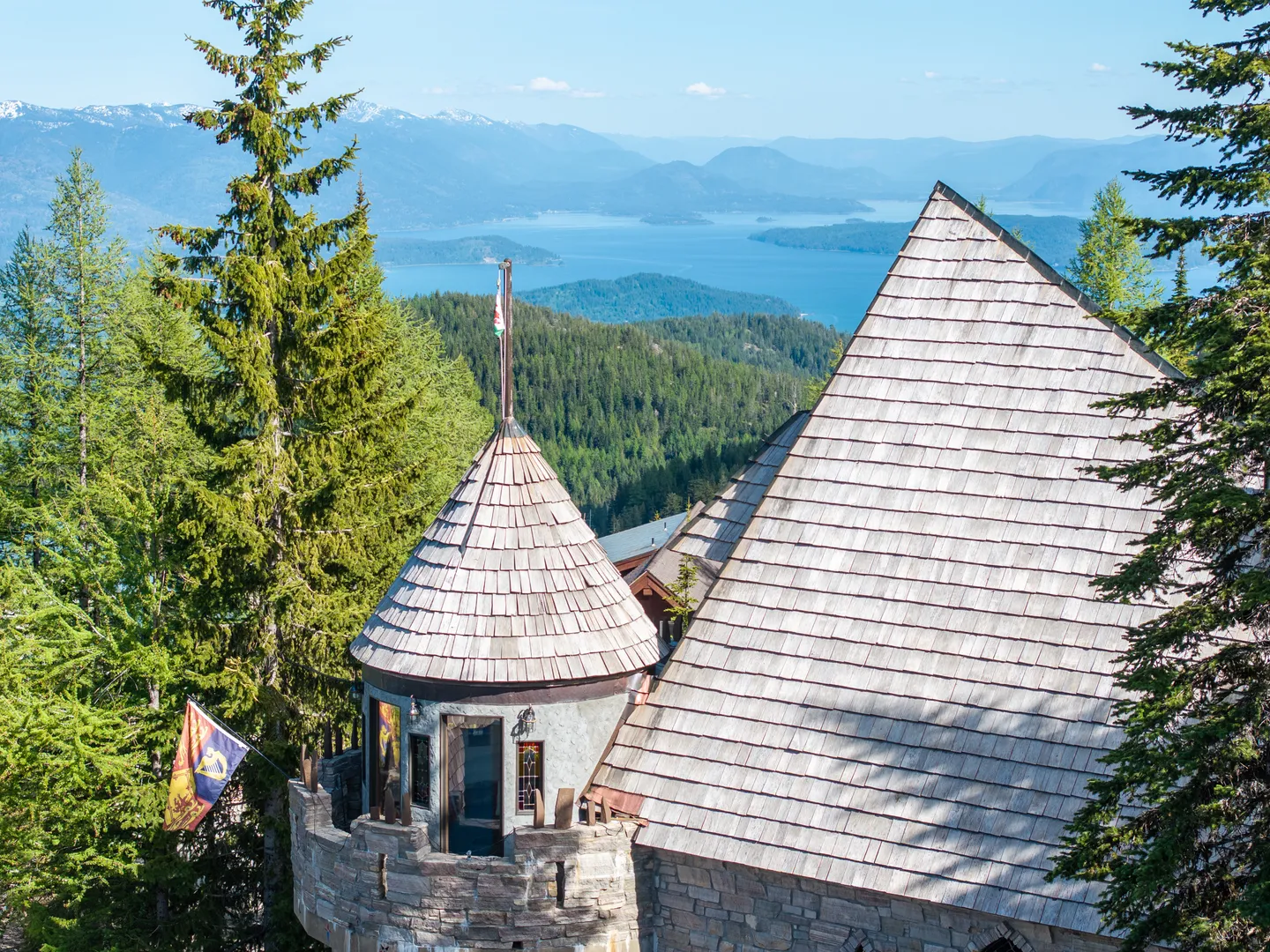 Exterior of a stone castle-style home with a conical roof tower overlooking forested mountains and a lake.