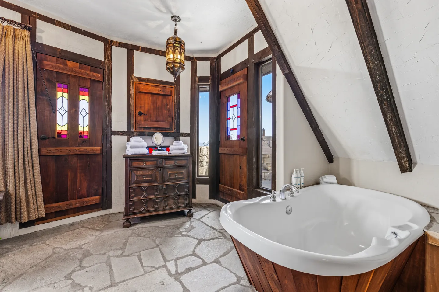 Bathroom with flagstone floors, a wood-paneled soaking tub, carved wooden dresser, and rustic doors with stained-glass inserts.