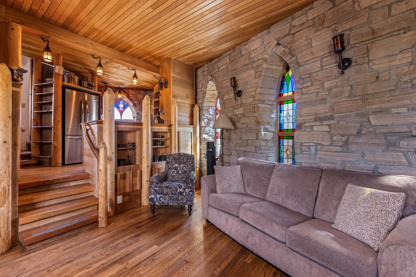 Living room featuring stone walls with arched stained-glass windows, wood-plank ceiling and floors, a couch, patterned armchair, and steps leading up to a kitchen area.