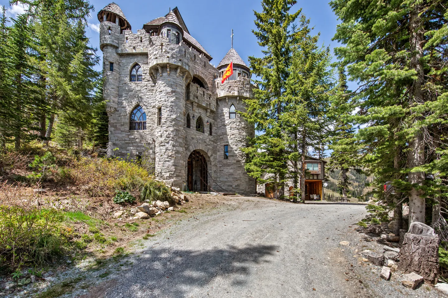 Exterior of a stone castle-style house with turrets, arched windows, and an arched entryway surrounded by pine trees under a blue sky.