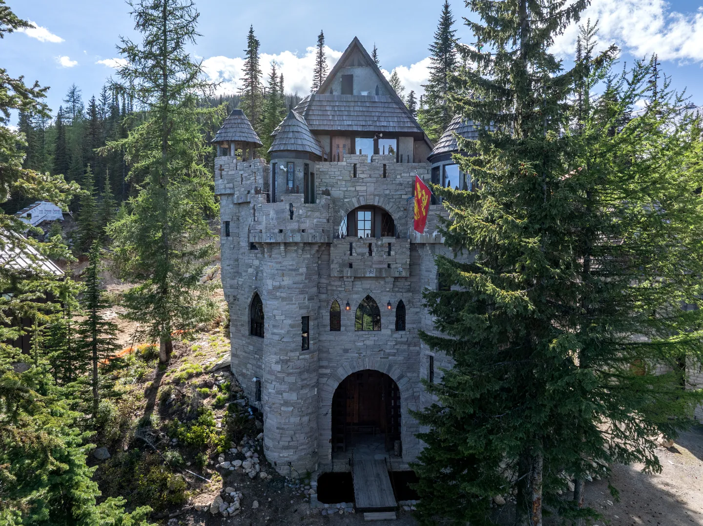 Exterior view of a stone castle-style house surrounded by tall pine trees, featuring turrets, an arched entry gate, and a red flag.
