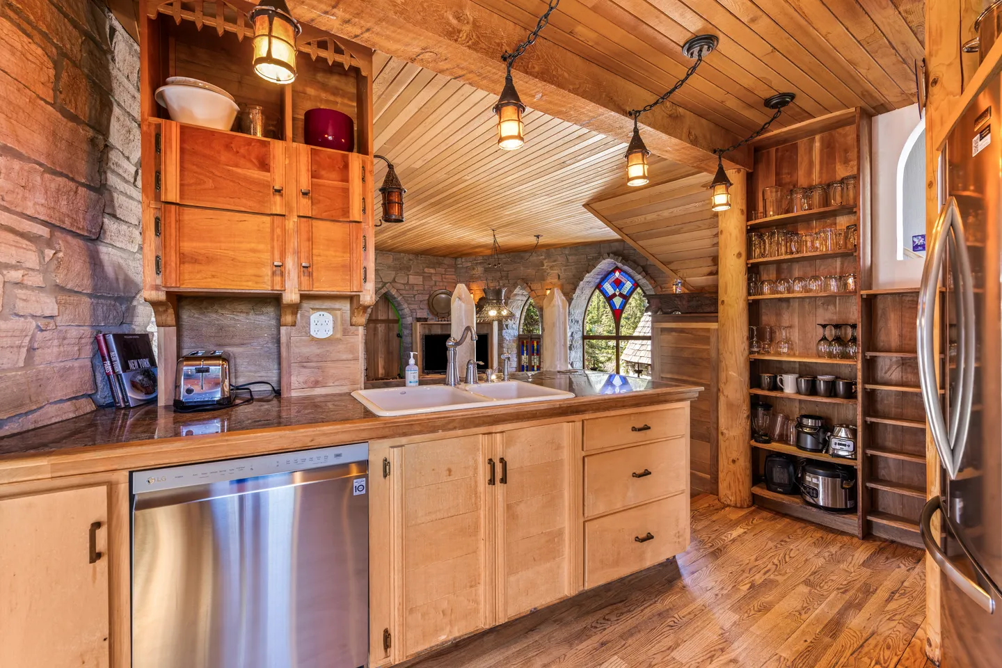 A rustic kitchen featuring wooden cabinets, a dishwasher, hanging pendant lights, and stone walls with arched stained-glass windows in the background.