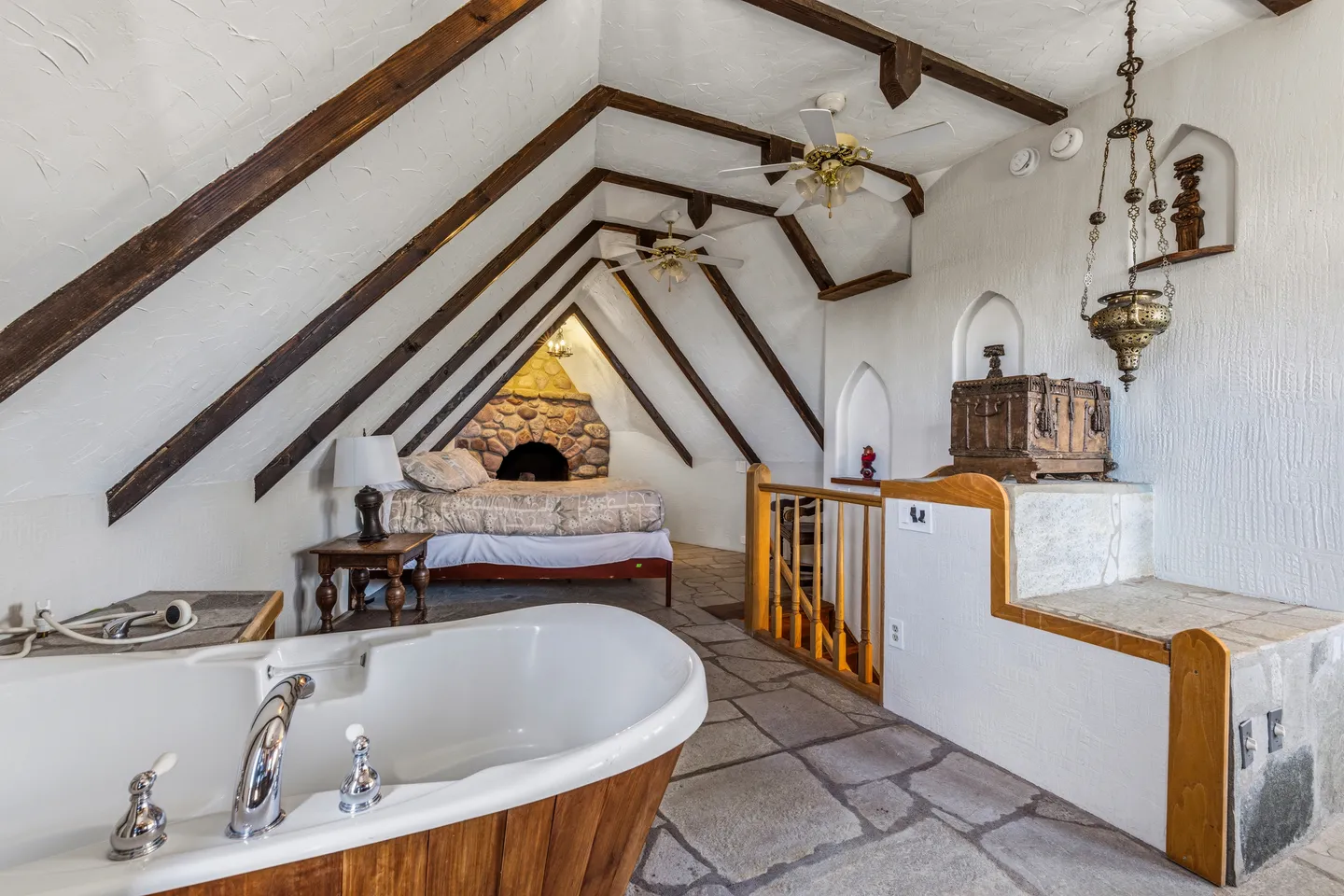 Attic-style bedroom with exposed ceiling beams, stone tile flooring, a soaking tub in the foreground, and a bed with a stone hearth in the background.