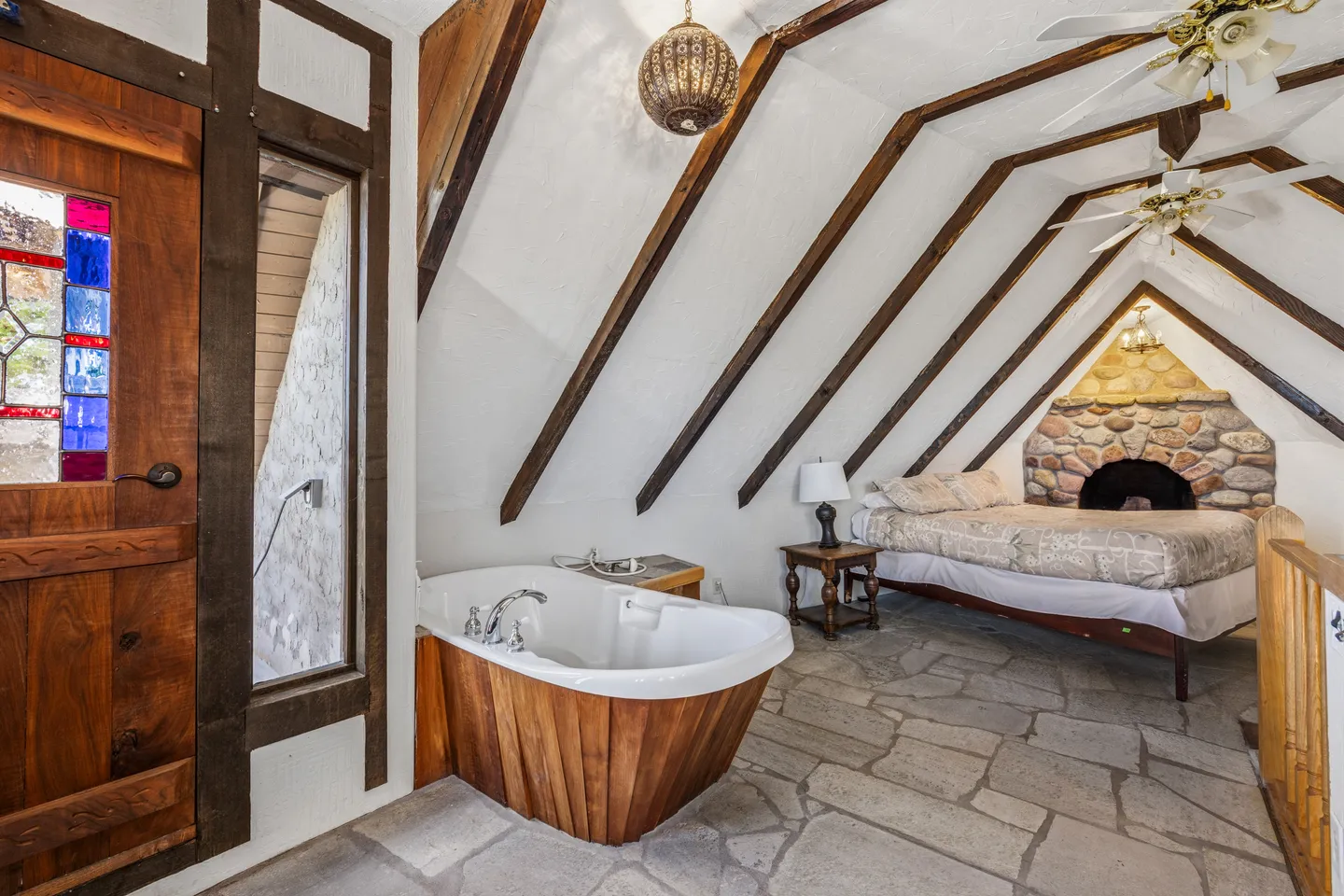 Attic bedroom with stone floor, pitched wood-beamed ceiling, soaking tub with wood paneling, and a bed near a stone fireplace.