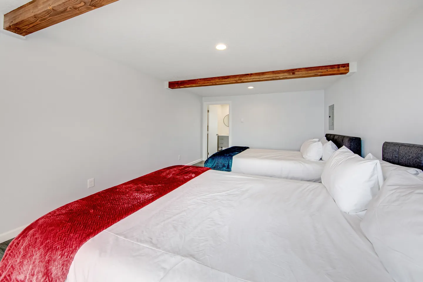 Bedroom featuring two beds made with white linens, one accented with a red throw blanket and the other with blue, beneath exposed ceiling beams.