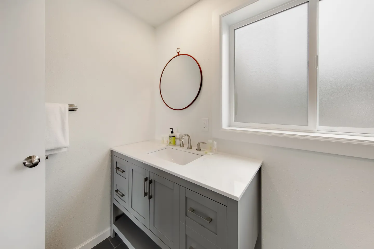 Bathroom vanity with a white countertop, undermount sink, gray base cabinets, and a round mirror beside a frosted window.
