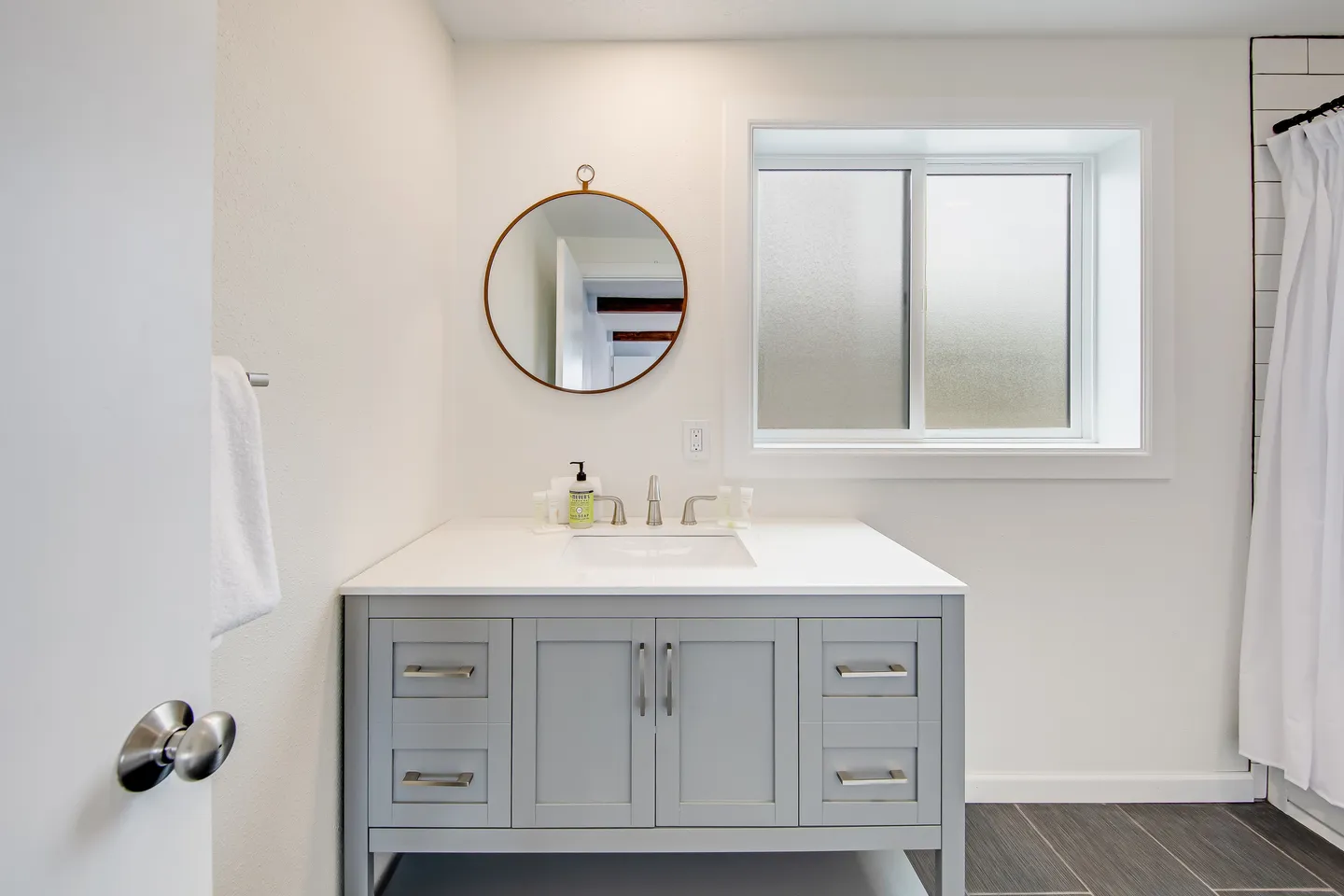 Bathroom vanity with a white countertop, grey cabinets, round mirror, and a frosted glass window.