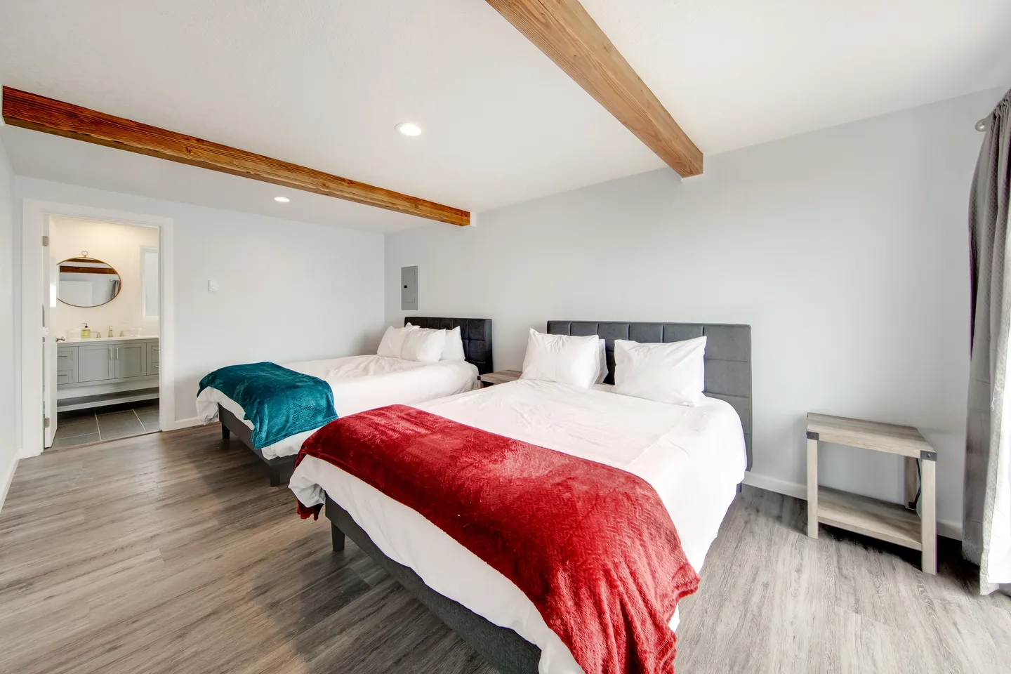 Bedroom with two beds, wood ceiling beams, wood-look flooring, and an open doorway leading to an ensuite bathroom.