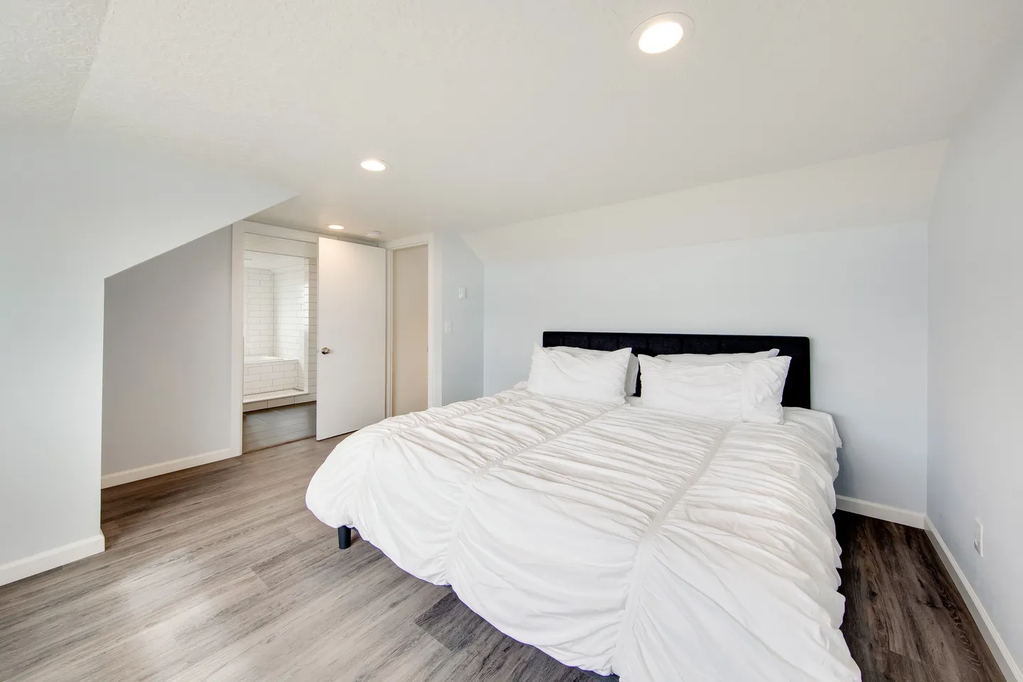 Bedroom with a white-linened bed, wood-look flooring, sloped ceilings, and an open door leading into an en-suite bathroom.