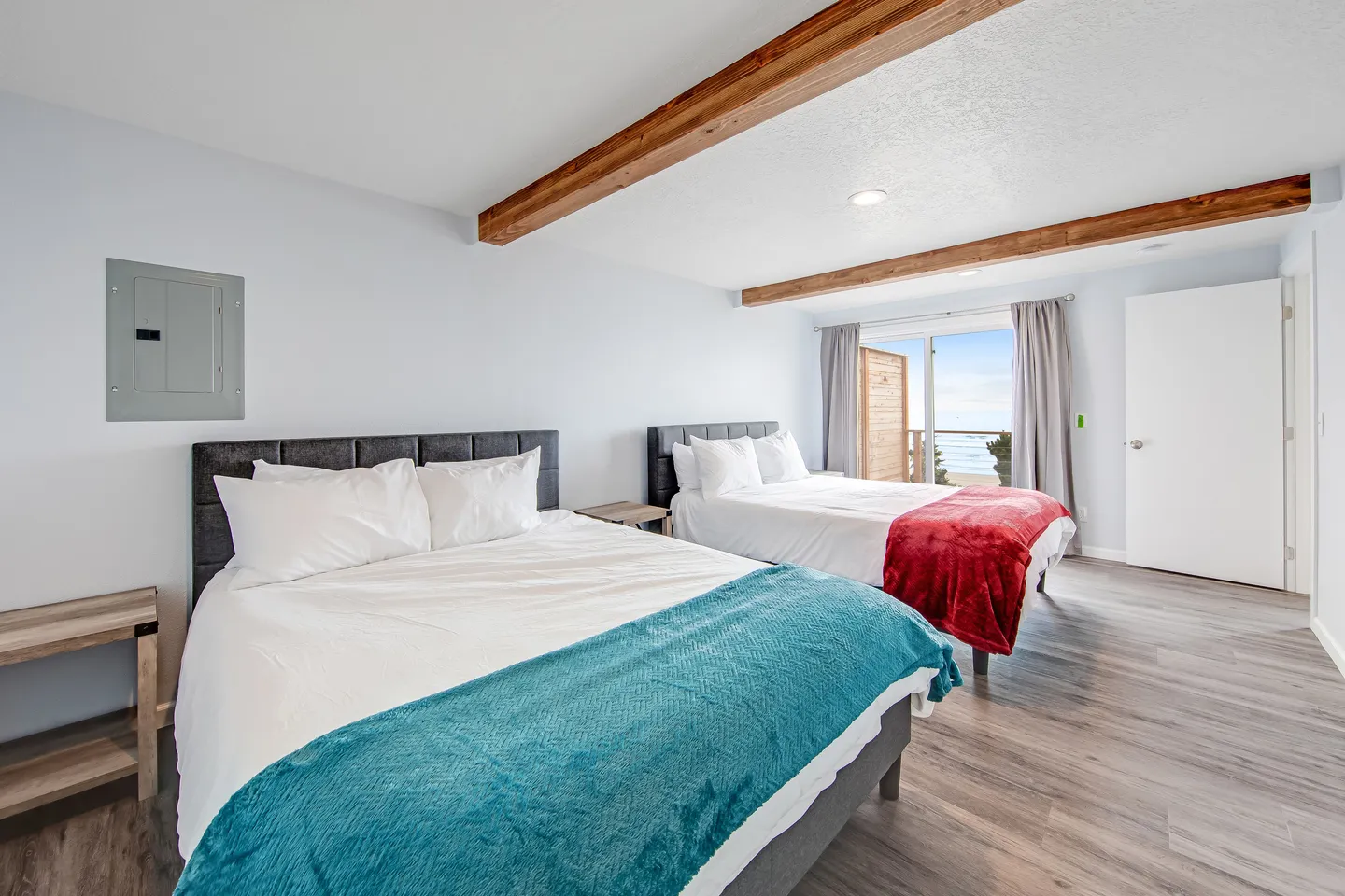 Bedroom with two beds, wood ceiling beams, wood-look flooring, and sliding glass doors leading to a balcony with an ocean view.